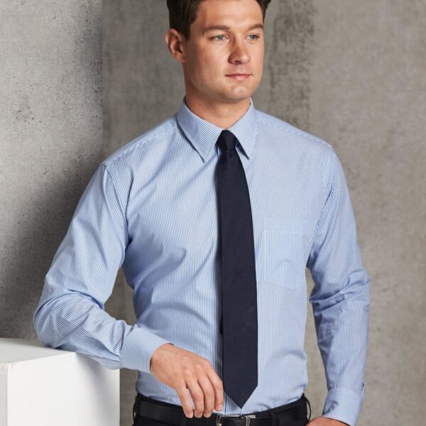 A man in a light blue, striped dress shirt and dark tie leans against a concrete wall. He stands with one hand resting on the surface, looking off to the side with a neutral expression. The background is a minimalist, industrial-style setting.
