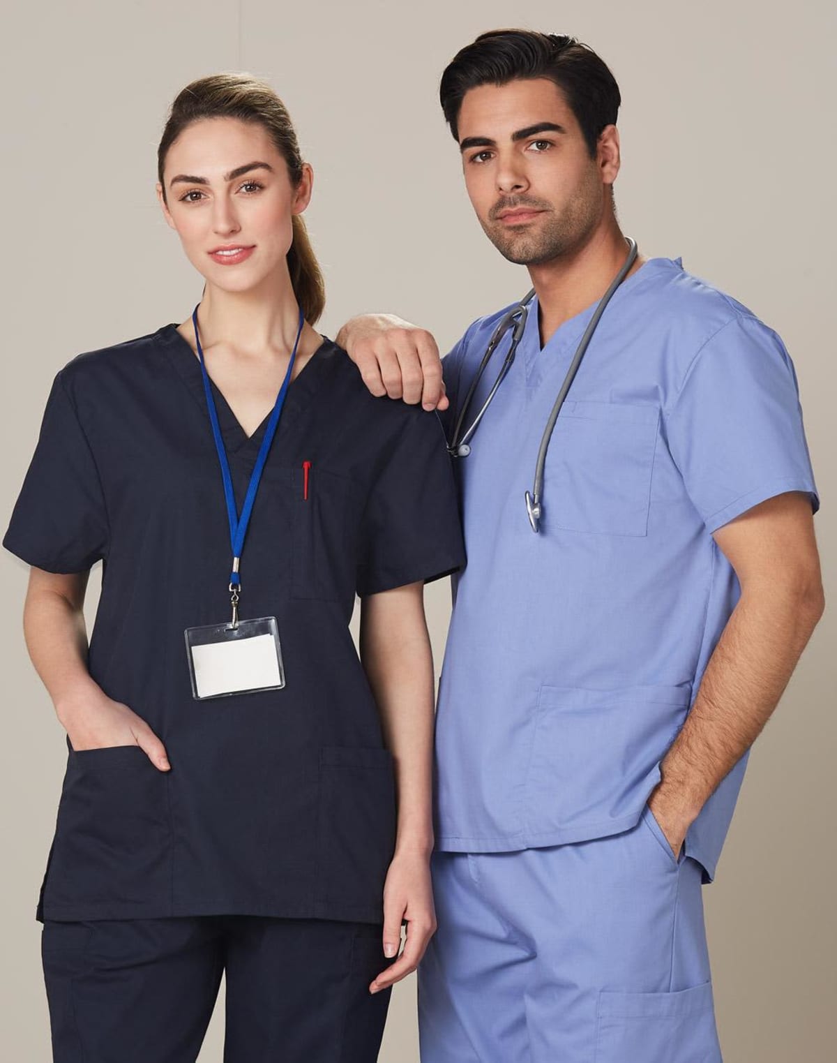 Two healthcare professionals in scrubs stand side by side. The woman on the left wears a dark scrub top with a nametag, and the man on the right wears a light blue scrub top with a stethoscope around his neck. Both look confidently at the camera.