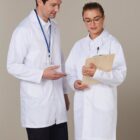 Two individuals wearing white lab coats stand together. One holds a clipboard while the other gestures towards it, engaged in conversation. Both wear black pants. The background is a neutral beige.