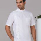 A man wearing a white chefs jacket with short sleeves and a high collar stands against a gray background. He is looking to the side and has his hand in his pocket. Theres a plant partially visible on the right.