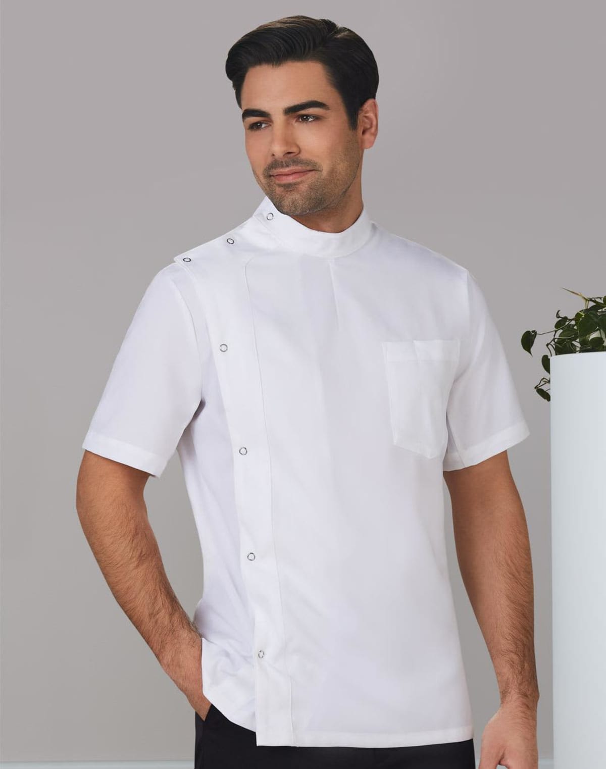 A man wearing a white chefs jacket with short sleeves and a high collar stands against a gray background. He is looking to the side and has his hand in his pocket. Theres a plant partially visible on the right.