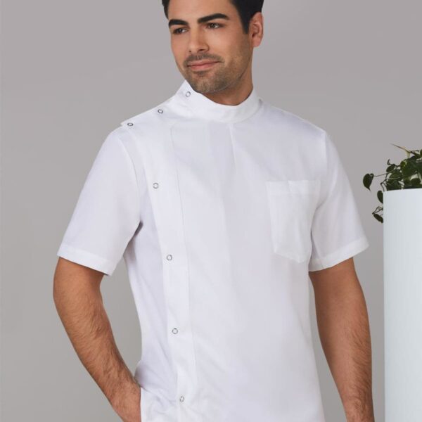 A man in a white short-sleeved chef jacket with a mandarin collar and snap buttons stands with his right hand in his pocket. He is looking to the side, and there is a plant in the background.