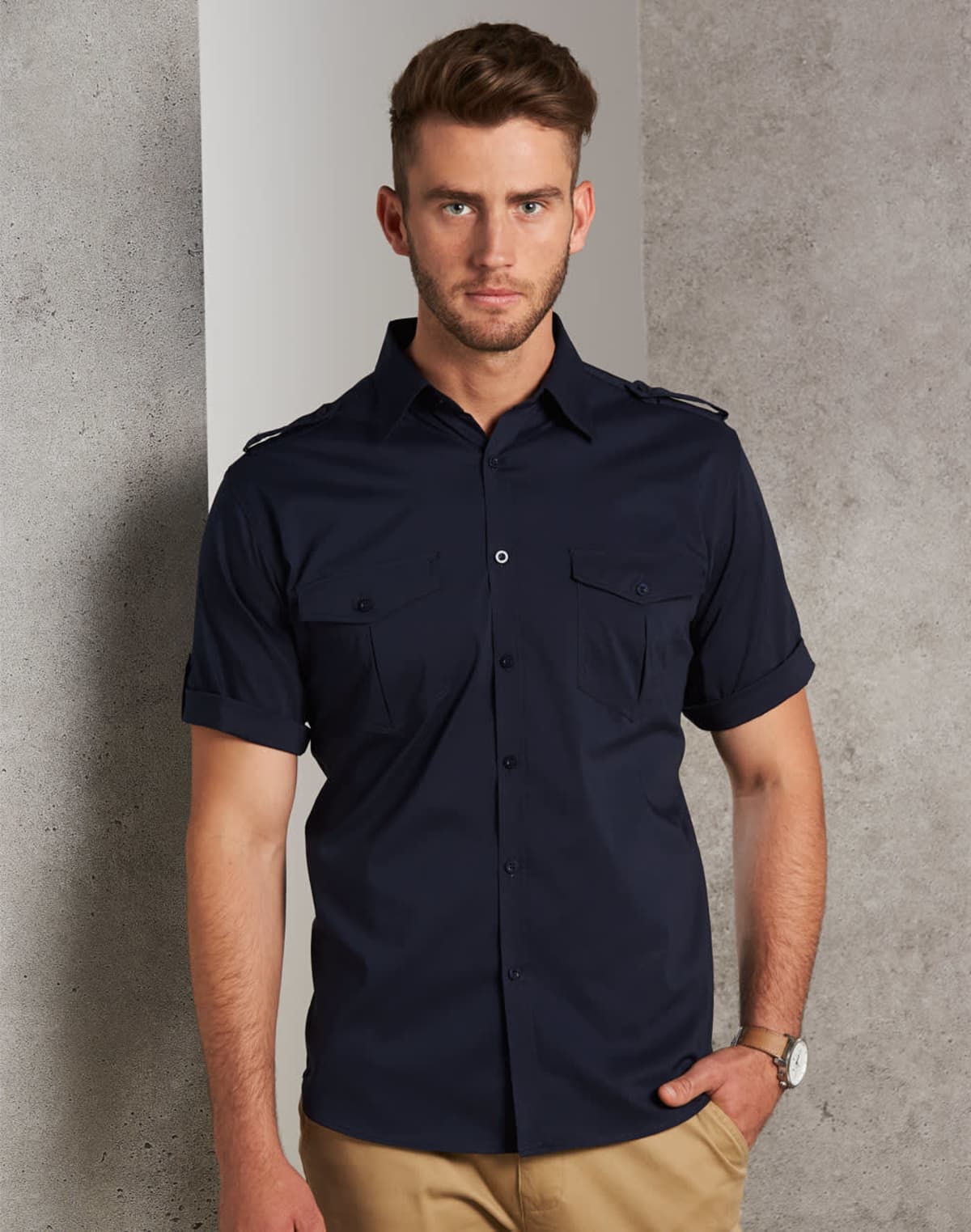 A man stands against a textured wall, wearing a navy short-sleeved button-up shirt with epaulettes and khaki pants. He has a watch on his left wrist and is looking directly at the camera.
