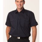 A man in a short-sleeve, dark blue button-up shirt with epaulets stands against a plain background. He is smiling, with his left hand in his pocket. The shirt has two chest pockets, and he wears a black belt.