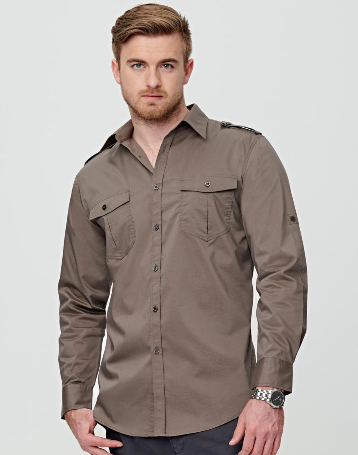A man with light brown hair and a beard wears a buttoned-up, long-sleeve, olive green shirt with chest pockets. He has his left hand in his pants pocket and stands against a plain light gray background.