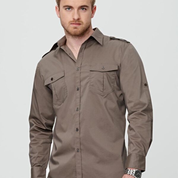 A man with light brown hair and a beard is wearing a gray button-up shirt with buttoned chest pockets. He poses slightly sideways with his right hand in his pocket, against a plain white background.