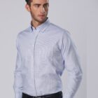 A person with short brown hair is wearing a light blue button-up shirt and dark pants. They stand with one hand in a pants pocket, looking to the side against a neutral gray background.