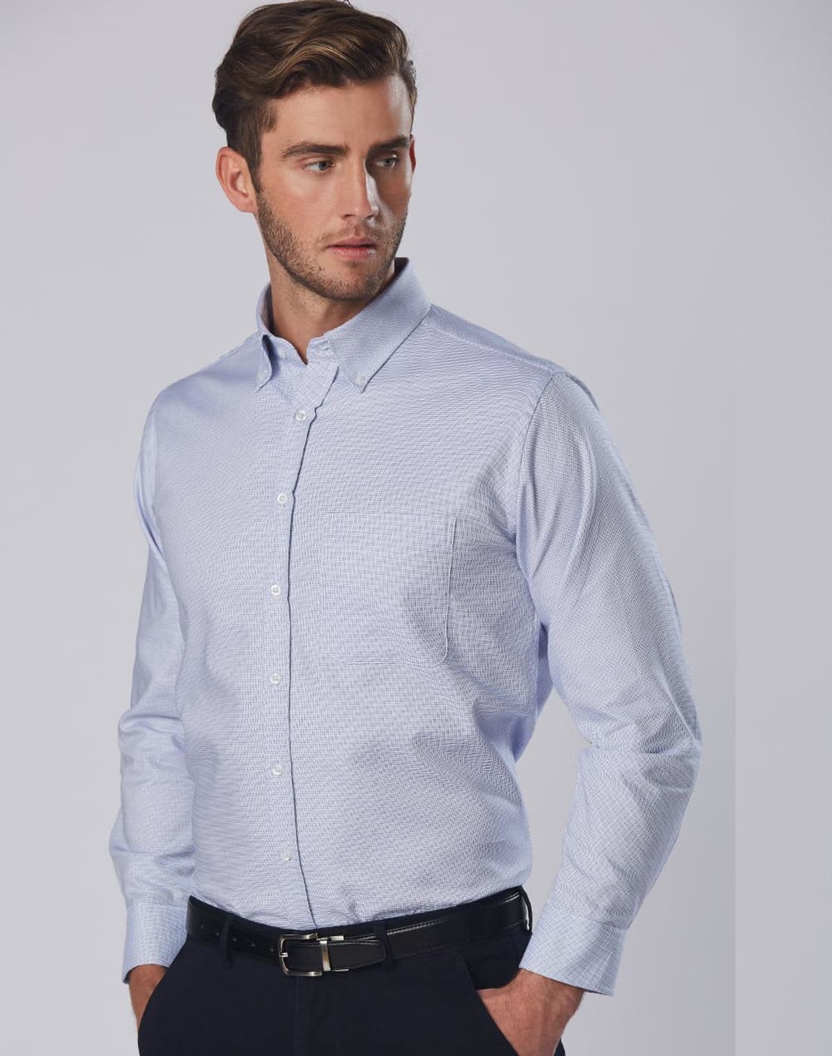 A person with short brown hair is wearing a light blue button-up shirt and dark pants. They stand with one hand in a pants pocket, looking to the side against a neutral gray background.