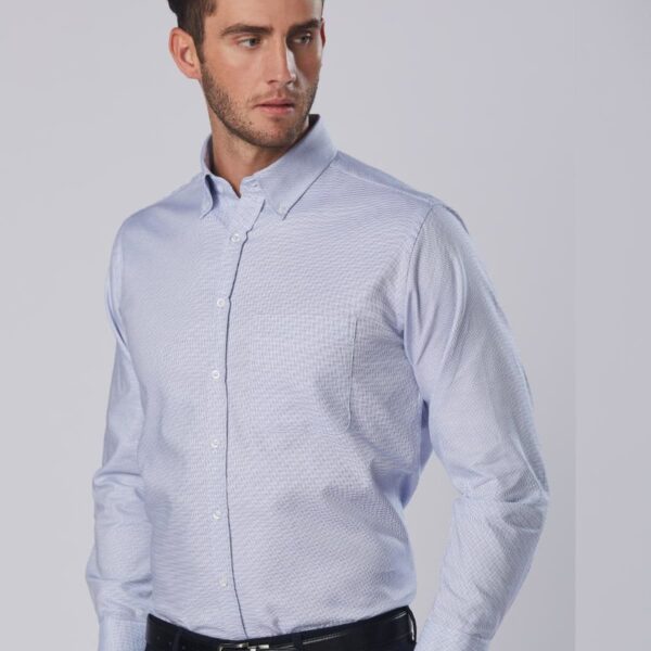 A person wearing a light blue button-up shirt looks to the side. They have short brown hair and a neatly groomed beard. The shirt is tucked into dark pants with a black belt. The background is plain and light-colored.