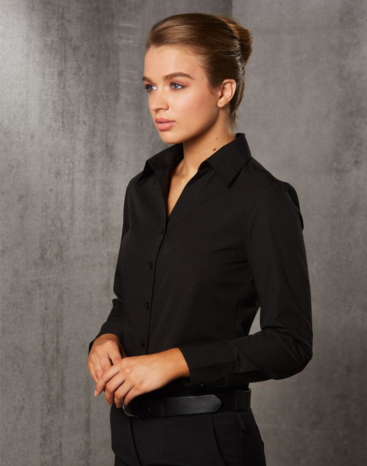A person with brown hair in a bun is wearing a black button-up shirt and black pants. They are standing against a gray textured wall, looking to the side with a neutral expression.