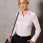 Person in a light pink blouse and black pants, holding a black bag, stands against a gray wall.