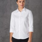 A person with tied-back hair is wearing a white long-sleeved button-up shirt and black pants, standing against a gray textured background.