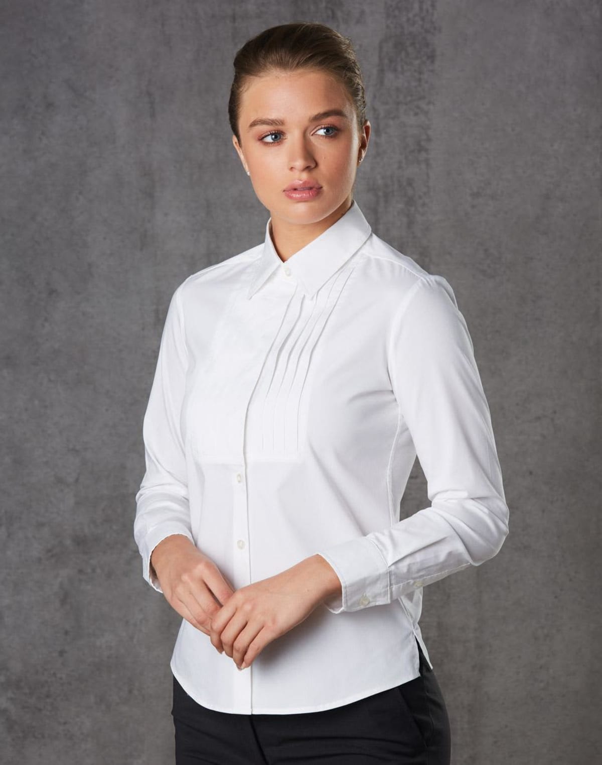 A person with brown hair is wearing a white, long-sleeved, button-up shirt with pleats on the chest. They stand against a gray background, looking to the side with a calm expression.
