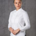 A person wearing a white button-down shirt with pleats on the chest stands against a gray textured background. The person is looking to the side with hands gently clasped in front.