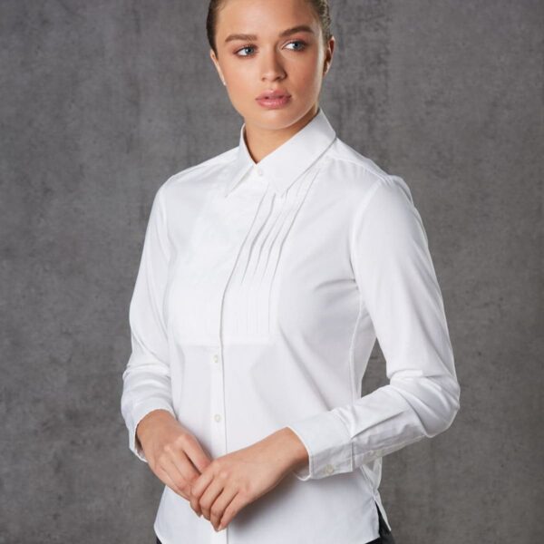 A person wearing a white button-down shirt with pleats on the chest stands against a gray textured background. The person is looking to the side with hands gently clasped in front.