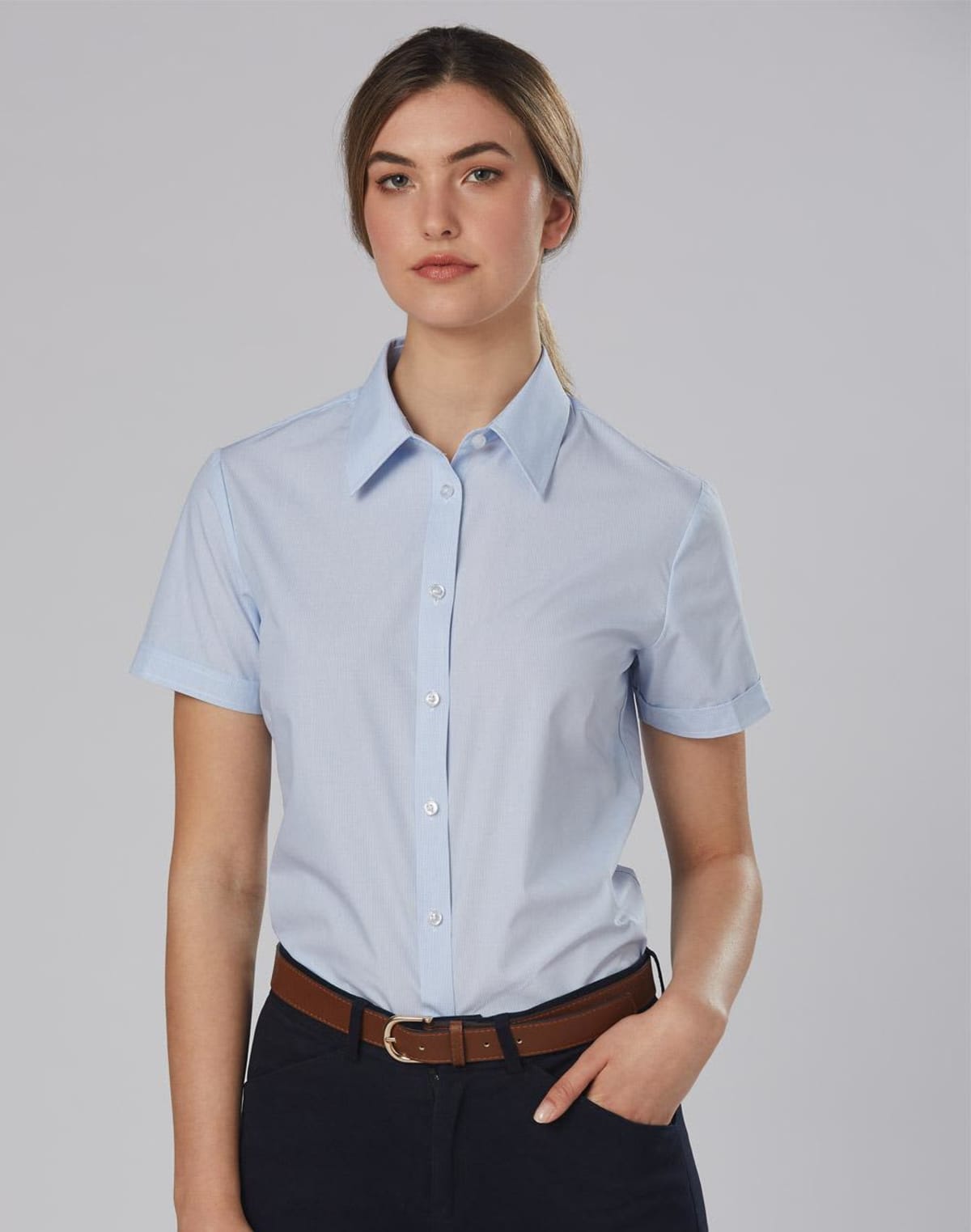 A person wearing a light blue short-sleeved button-up shirt and dark pants stands with one hand in their pocket. They have tied-back hair and are set against a neutral gray background.