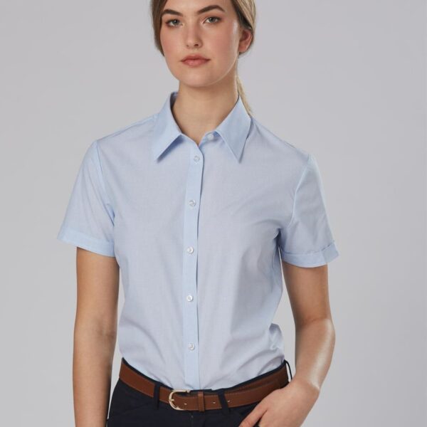 A person wearing a light blue short-sleeved button-up shirt and dark pants stands against a plain gray background. They have their left hand in their pocket and a neutral expression.