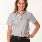 Woman smiling, wearing a light gray short-sleeved button-up shirt and black pants. She has her right hand on her hip and is standing against a plain white background.