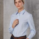 Person wearing a light blue button-up shirt and dark pants stands against a textured gray background. They have their hair pulled back and are gently holding their own collar with one hand.