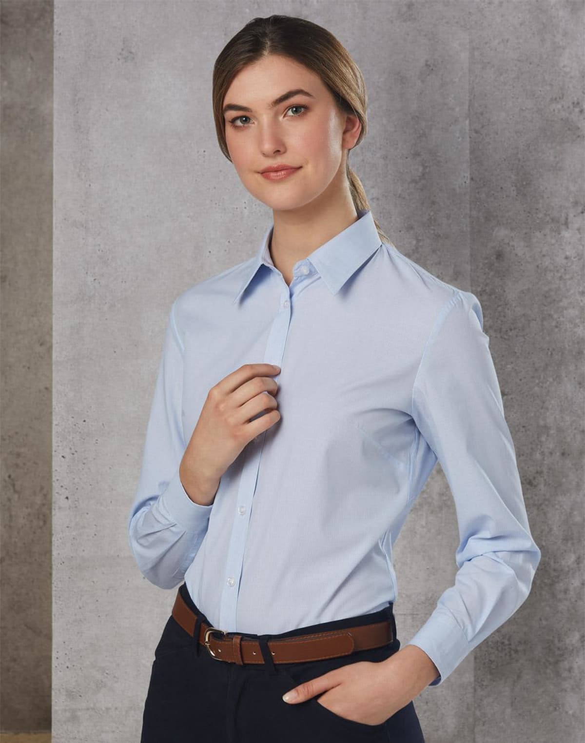 Person wearing a light blue button-up shirt and dark pants stands against a textured gray background. They have their hair pulled back and are gently holding their own collar with one hand.