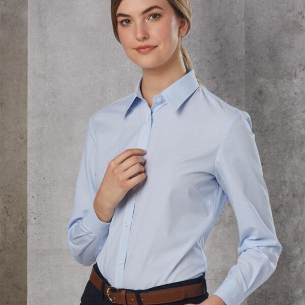 A person stands confidently against a gray textured wall, wearing a light blue button-up shirt and dark pants with a brown belt. They have their hair pulled back and hold one hand near their collar.
