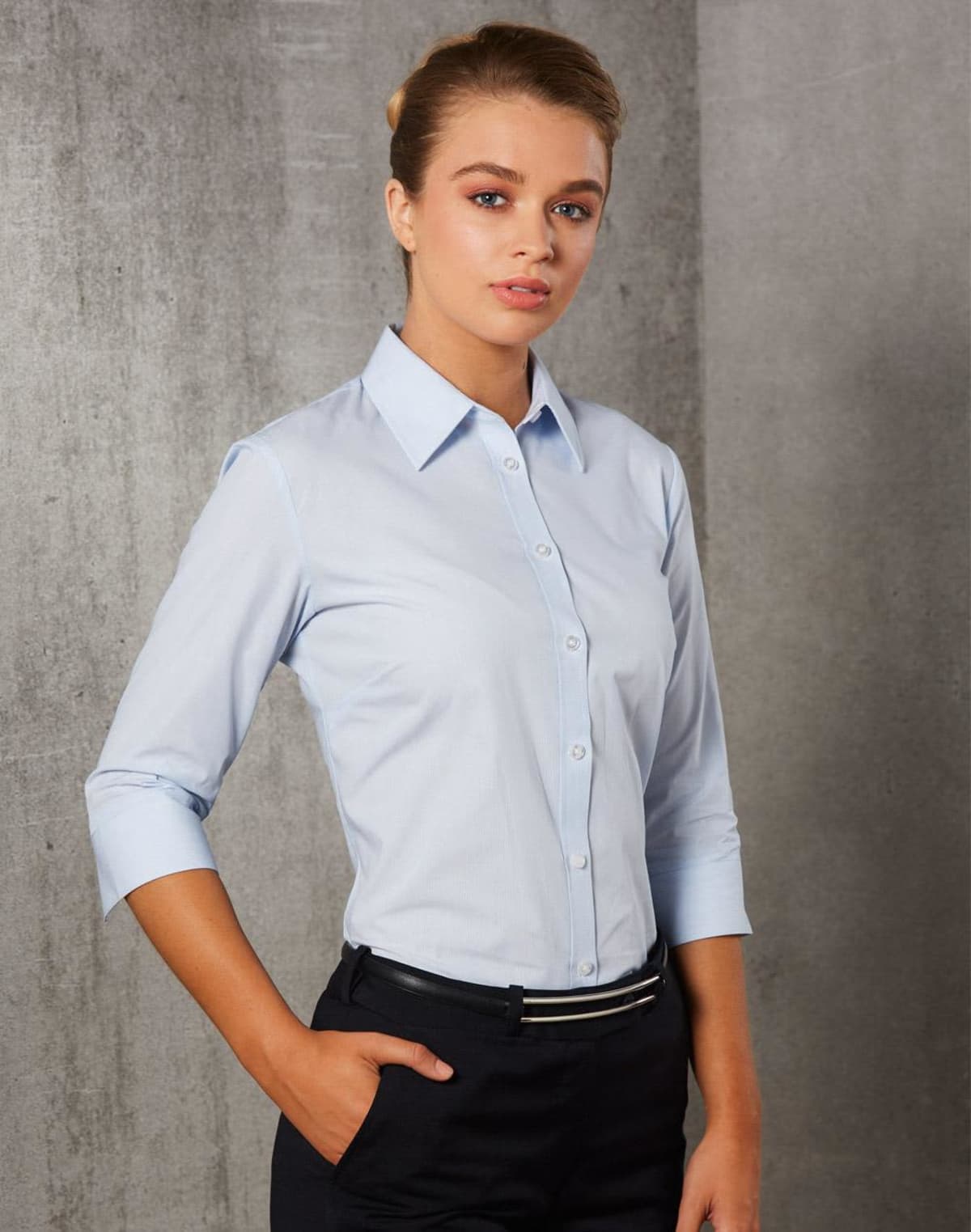 Person wearing a light blue button-down shirt and black pants stands against a gray textured background. They have their hair neatly tied back and are looking confidently at the camera.