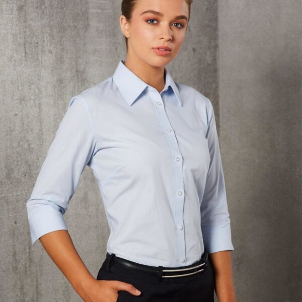 A person stands against a gray textured background, wearing a light blue button-up shirt with three-quarter sleeves and dark pants. The persons hair is neatly styled, and they have a confident expression.