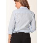 A woman with brown hair is standing with her back to the camera, wearing a light blue button-up shirt with rolled-up sleeves and black pants.
