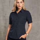 A woman with shoulder-length blonde hair is wearing a dark pinstripe shirt and dark pants. She is looking slightly to the side with one hand in her pocket, standing against a plain gray background.