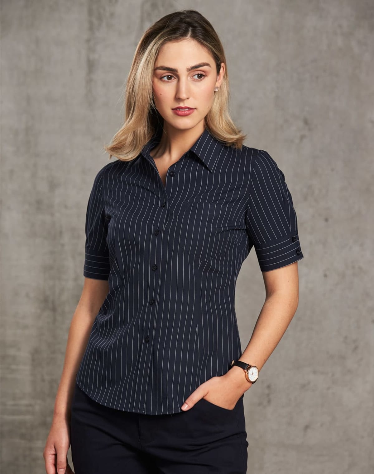 A woman with shoulder-length blonde hair is wearing a dark pinstripe shirt and dark pants. She is looking slightly to the side with one hand in her pocket, standing against a plain gray background.
