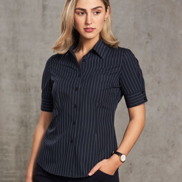 A person with shoulder-length blonde hair is wearing a dark, short-sleeved, pinstripe button-up shirt and dark pants, standing against a plain gray background. One hand rests in a pocket, and the other is at their side.