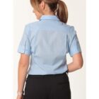 Woman with brown hair in a ponytail, wearing a light blue short-sleeve button-up shirt and black pants, standing with her back facing the camera against a plain white background.