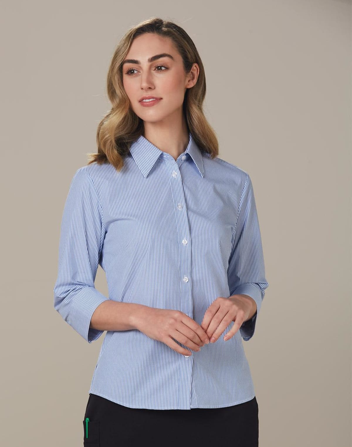 A woman with shoulder-length hair wears a light blue, pinstripe, button-up blouse. The blouse has three-quarter sleeves. She stands against a plain, neutral background with hands gently clasped.