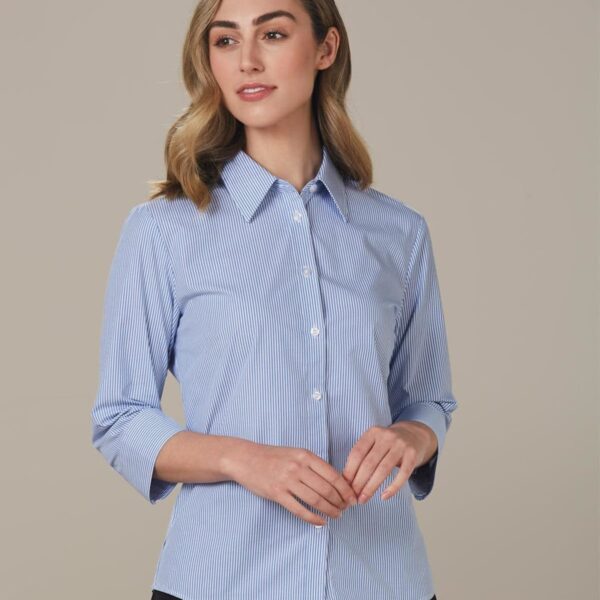 A person with wavy blonde hair is wearing a blue and white striped button-up shirt with three-quarter sleeves. They stand against a neutral background, looking slightly to the side with hands gently clasped.