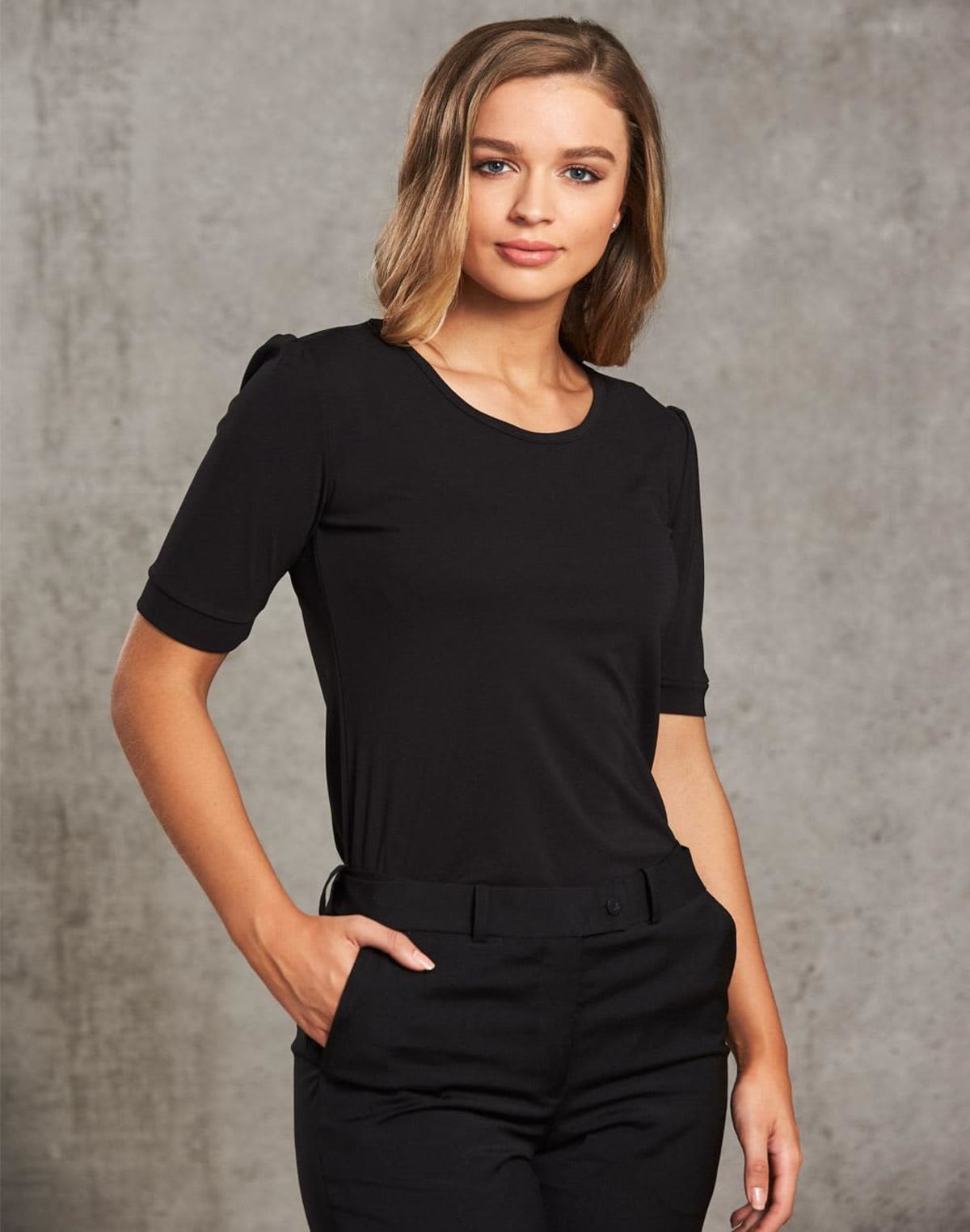 A person with shoulder-length hair wearing a black, short-sleeved shirt and pants stands with one hand in their pocket against a textured gray background. They are looking directly at the camera with a neutral expression.