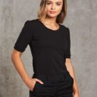 A person with shoulder-length hair is wearing a black t-shirt and black pants against a gray textured background. They have their left hand in their pocket and are looking at the camera.