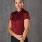 A woman stands against a gray background, wearing a burgundy blouse and black pants. She has her hair tied back and is looking directly at the camera with a slight smile, her hands gently clasped in front of her.