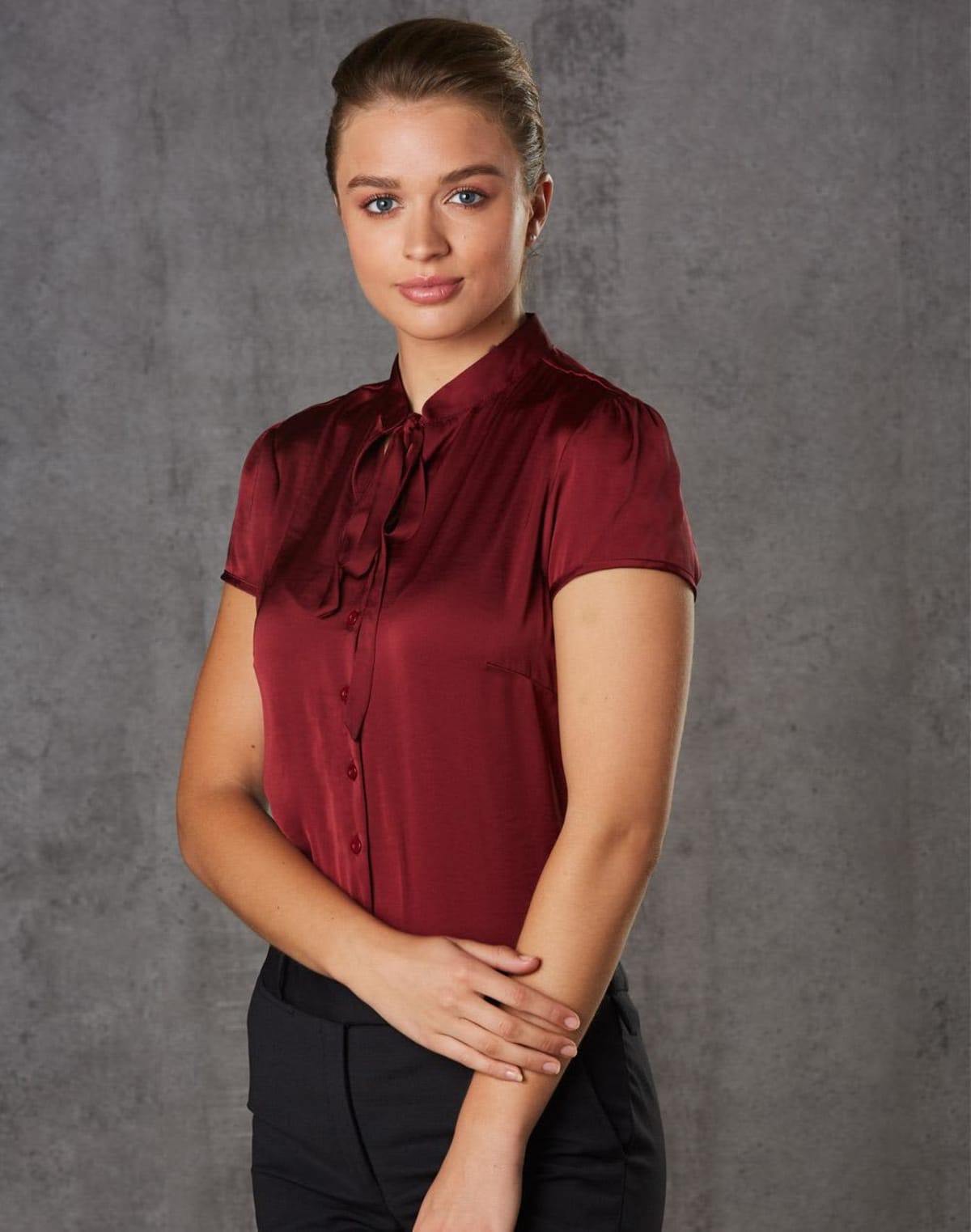 A woman stands against a gray background, wearing a burgundy blouse and black pants. She has her hair tied back and is looking directly at the camera with a slight smile, her hands gently clasped in front of her.