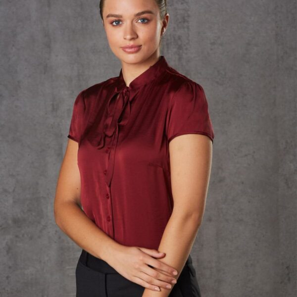 A woman stands in front of a gray textured background. She is wearing a burgundy blouse with a bow and black pants. She has her hair pulled back and is smiling softly while looking at the camera, with her hands gently crossed in front.