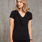 A woman with shoulder-length blonde hair is wearing a black short-sleeve top and black pants. She stands against a gray concrete wall, looking to her right with a neutral expression. The top features a ruffled V-neck design.