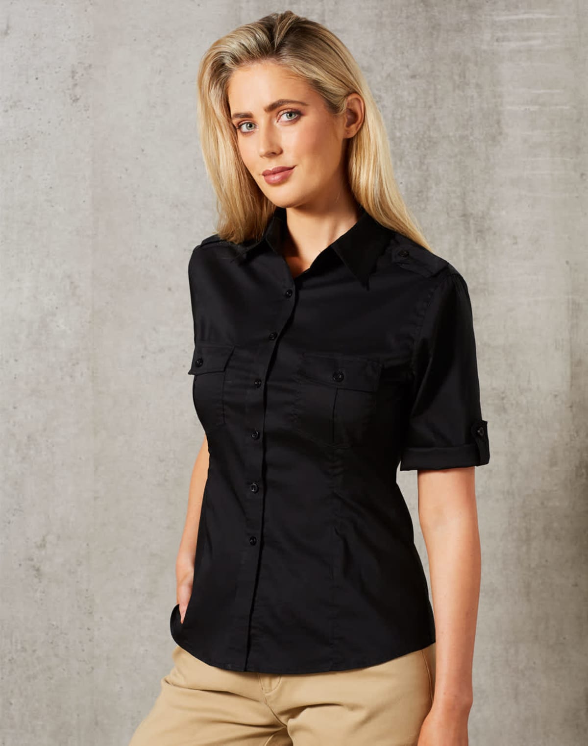 A person with long blonde hair wearing a black button-up shirt and beige pants stands against a gray textured background. They have a neutral expression and hands in their pockets.