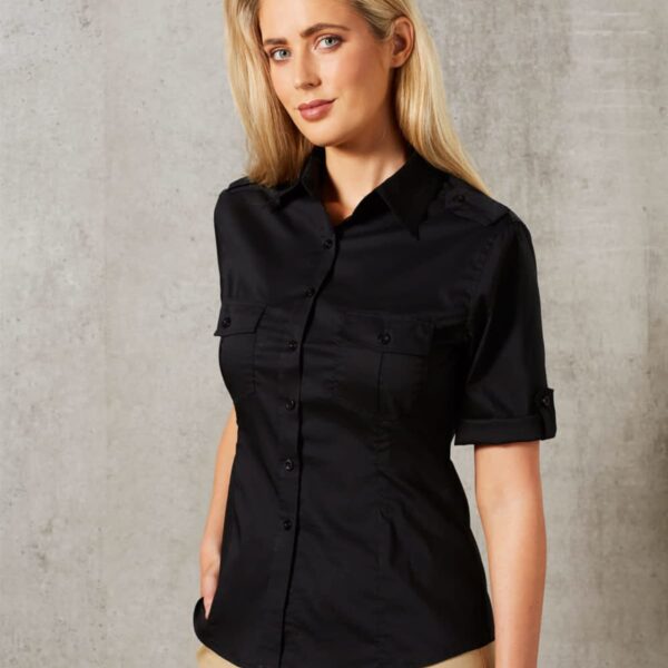 A woman with long blonde hair is wearing a black button-up shirt and beige pants. She is standing against a textured gray background with one hand in her pocket, looking at the camera.