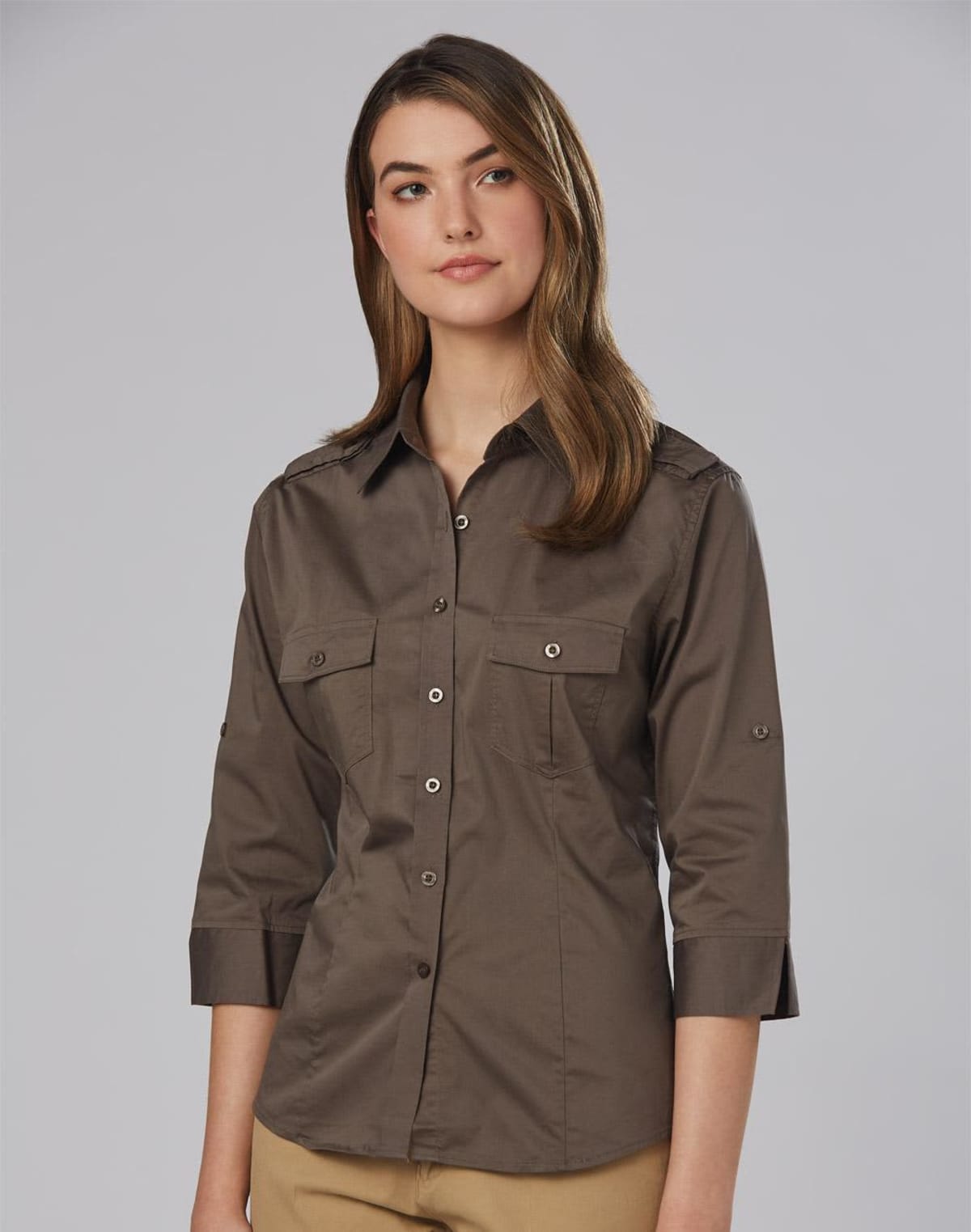 A person with long hair wearing a brown button-up shirt with three-quarter sleeves stands against a plain gray background, looking slightly to the side with a neutral expression.