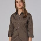 A person with long brown hair is wearing a dark khaki button-up shirt with rolled-up sleeves and beige pants. The shirt has two chest pockets with flap closures and buttons. The background is a plain light gray.