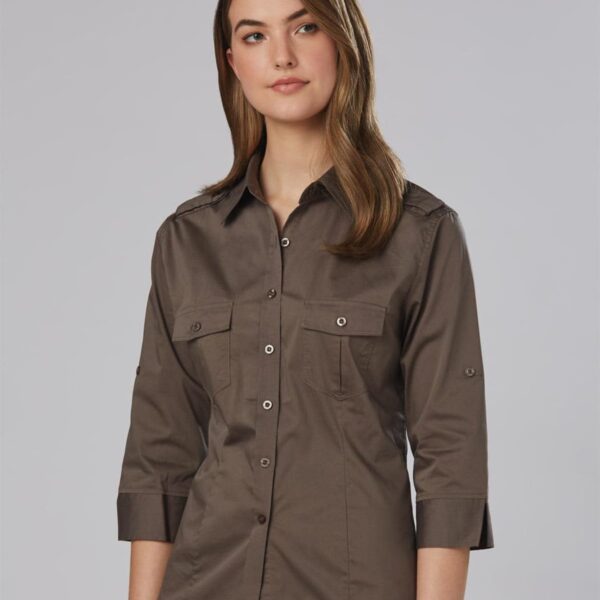 A person with long brown hair is wearing a dark khaki button-up shirt with rolled-up sleeves and beige pants. The shirt has two chest pockets with flap closures and buttons. The background is a plain light gray.