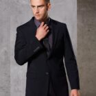 Man in a dark suit and black shirt adjusting his tie. He is posed against a plain, gray background, looking off to the side.