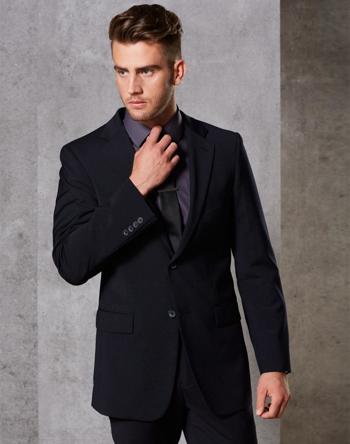 Man in a dark suit and black shirt adjusting his tie. He is posed against a plain, gray background, looking off to the side.