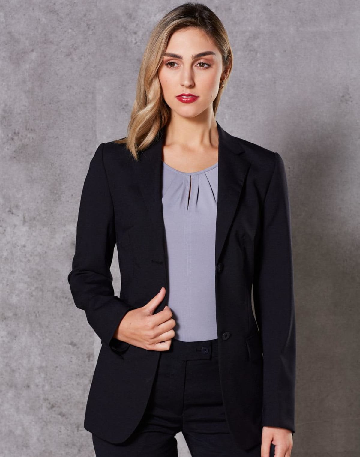 A woman with light brown hair wearing a black blazer over a gray blouse stands against a gray textured background. She gazes slightly to the side, with a neutral expression, and holds one side of her blazer with her hand.
