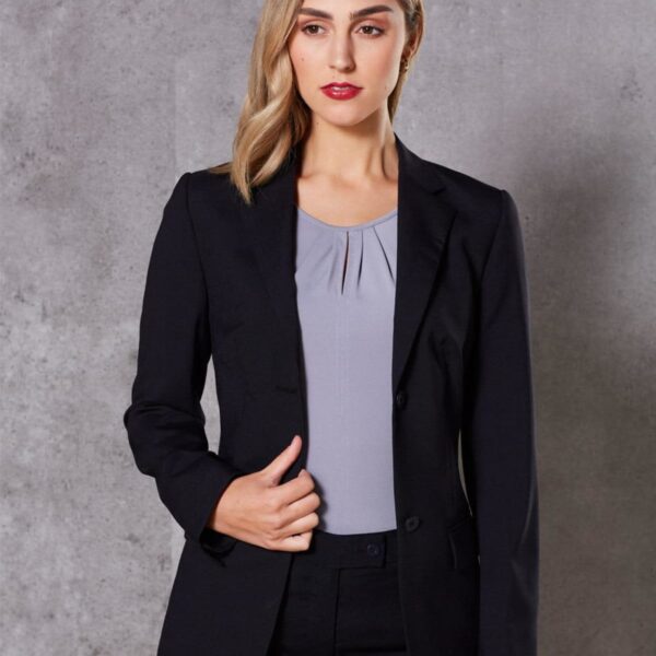 A person with long hair, wearing a dark blazer over a light blouse, stands against a gray textured wall. They have one hand holding the blazer lapel and are looking slightly to the side.