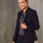 A woman in a dark blazer and shirt smiles while standing against a gray, textured wall. Her hair is neatly tied back, and she holds the lapel of her blazer with one hand.